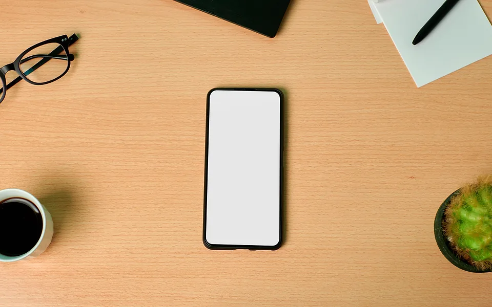 Top-down view of a phone with a blank white screen on a timber desk beside glasses, coffee and a cactus