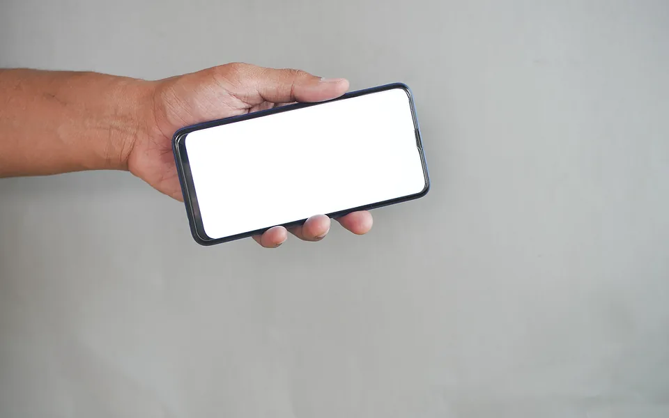 Hand holding a smartphone sideways with a blank white screen against a plain grey wall