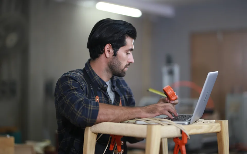 Tradie in a checked shirt typing on a laptop set on a workshop stool, an orange handheld tool in his other hand