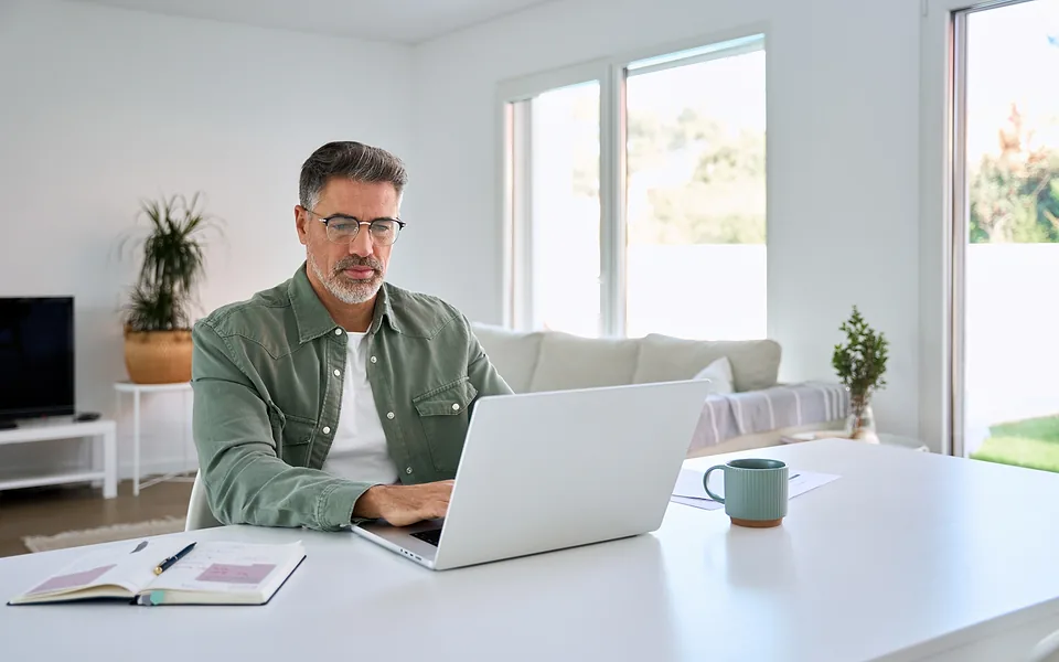 Grey-haired man in glasses and a green shirt using a laptop at a white table in a bright living room