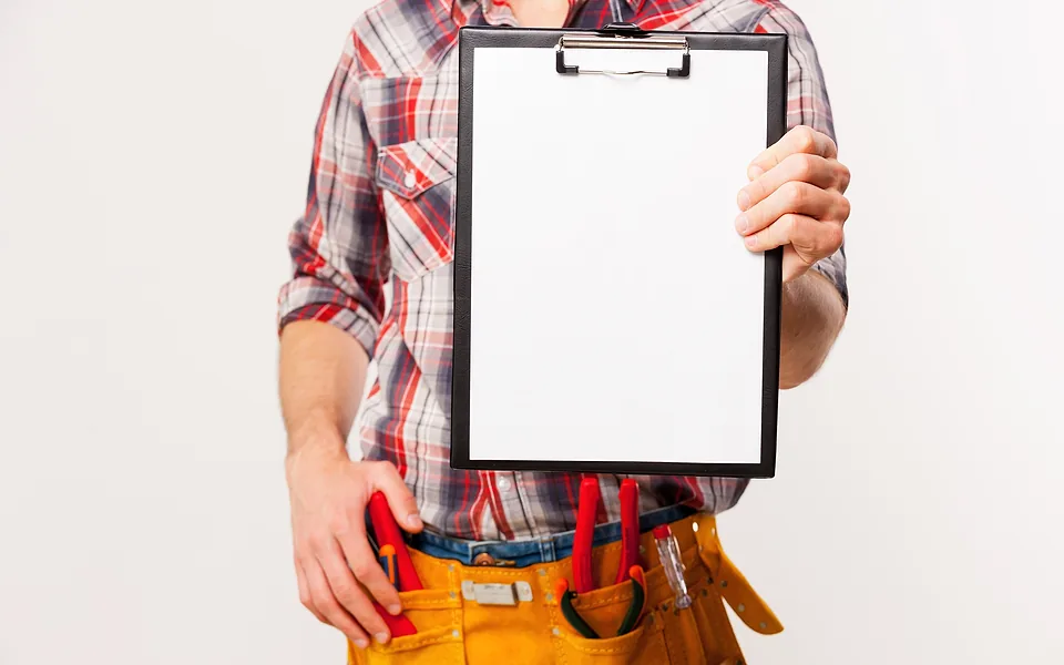 Tradesman in a checked shirt and tool belt holding up a clipboard with a blank white sheet of paper