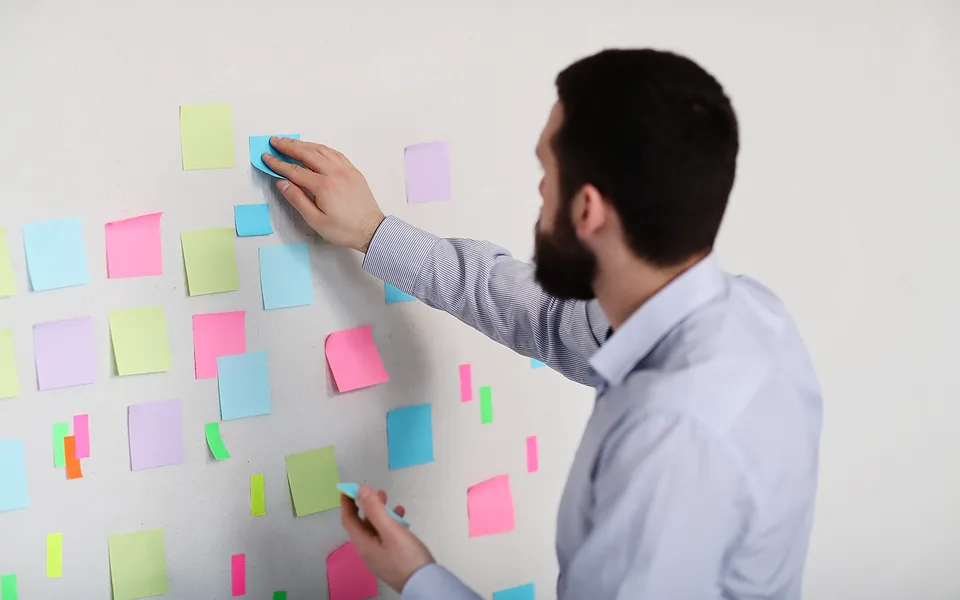 Bearded man in a business shirt arranging rows of colourful sticky notes across a white wall