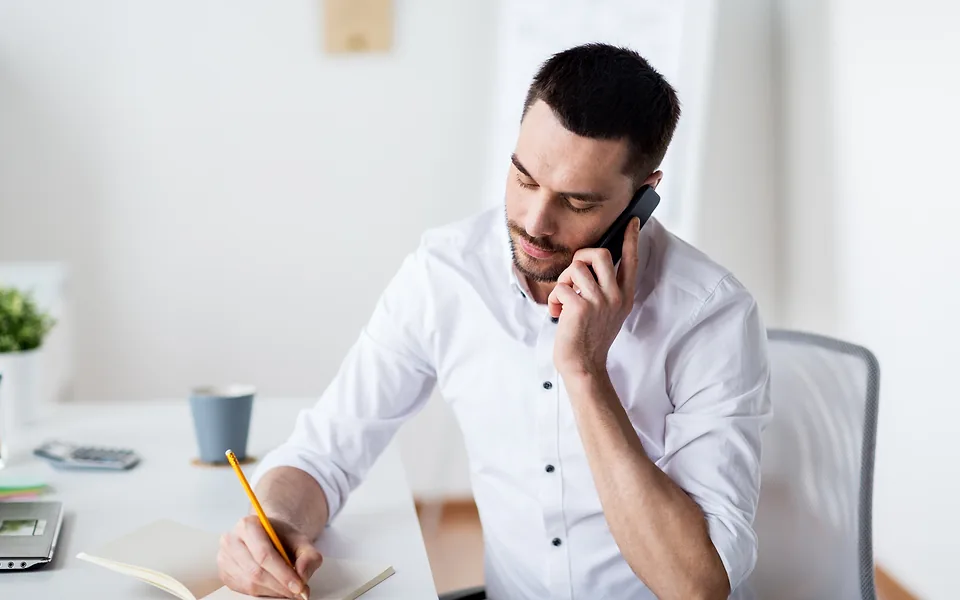 Man in a white shirt talking on a mobile phone while writing notes with a pencil at his desk