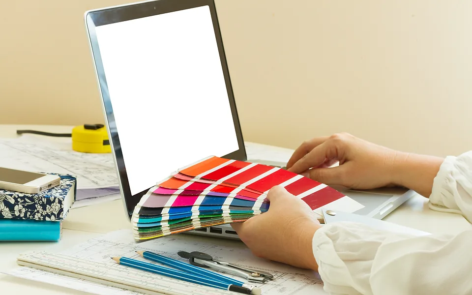 Hands fanning out a deck of colour swatches in front of a laptop with a blank white screen