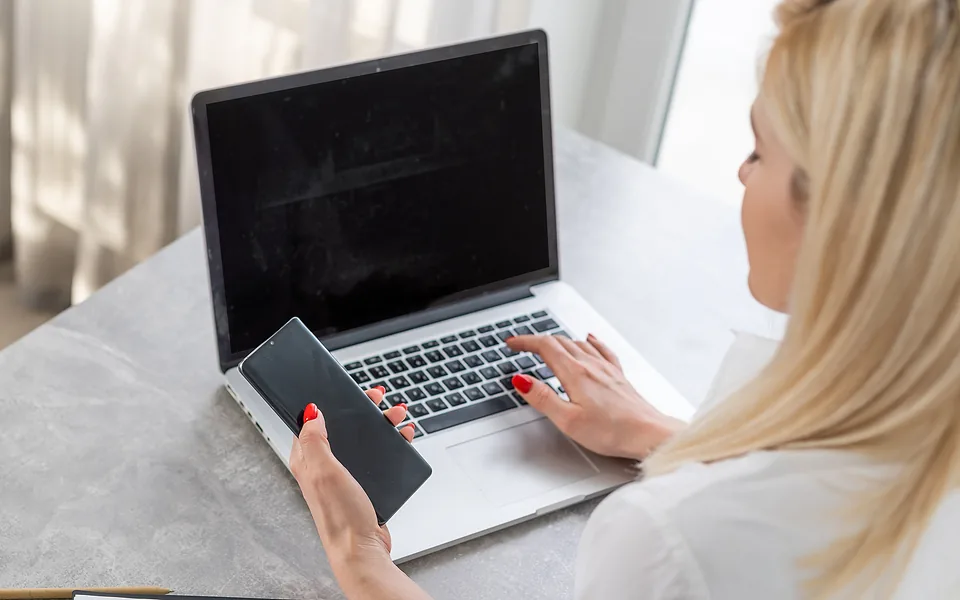 Blonde woman typing on a laptop with one hand while holding a mobile phone in the other
