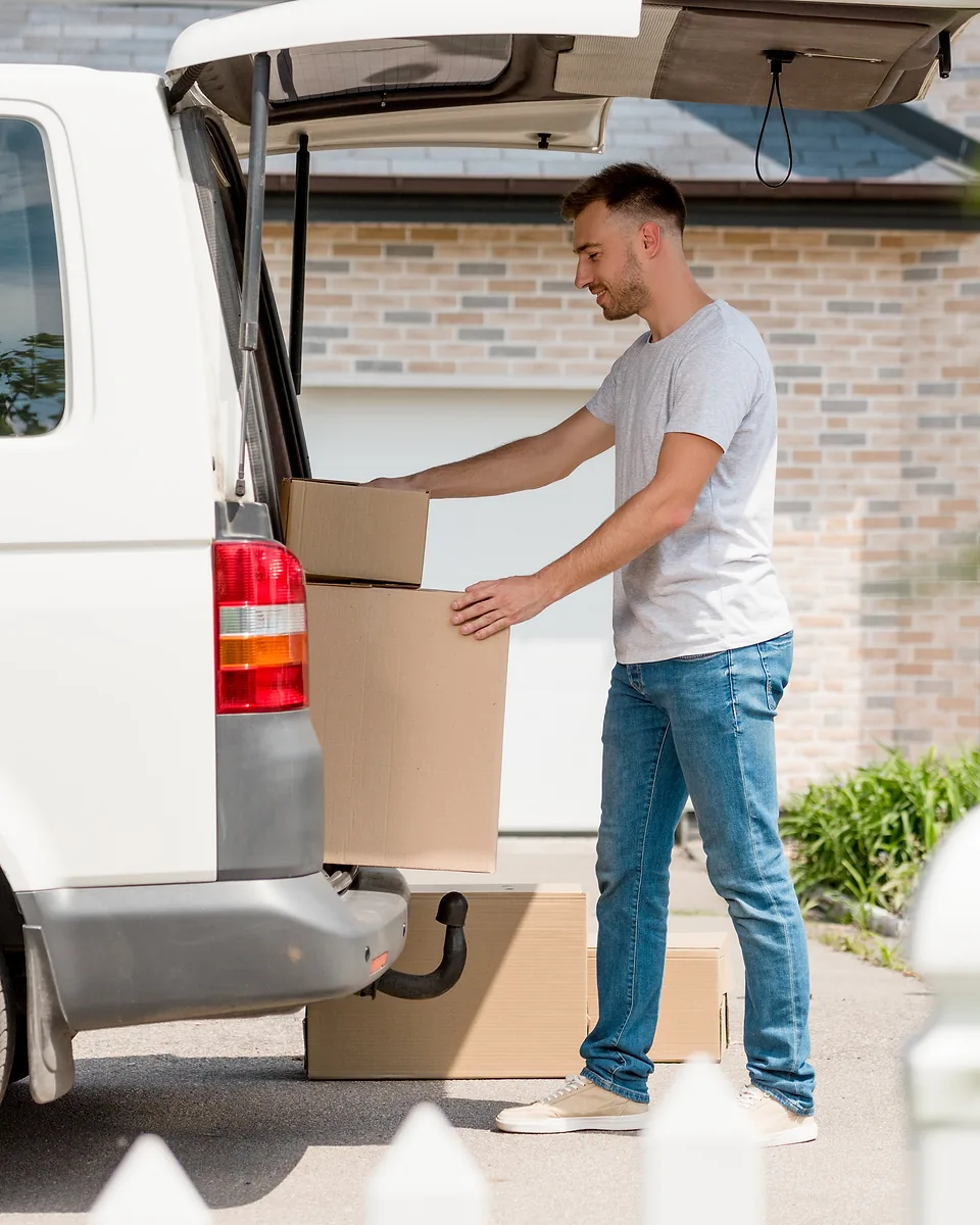 Man in a grey t-shirt unloading cardboard boxes from the back of a white van outside a brick house