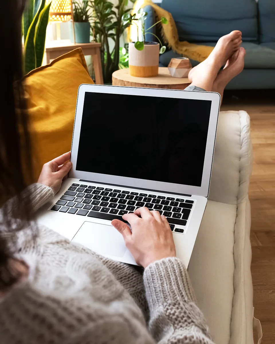 Working remotely from a sofa at home, a person in a knitted jumper types on a laptop resting on their lap