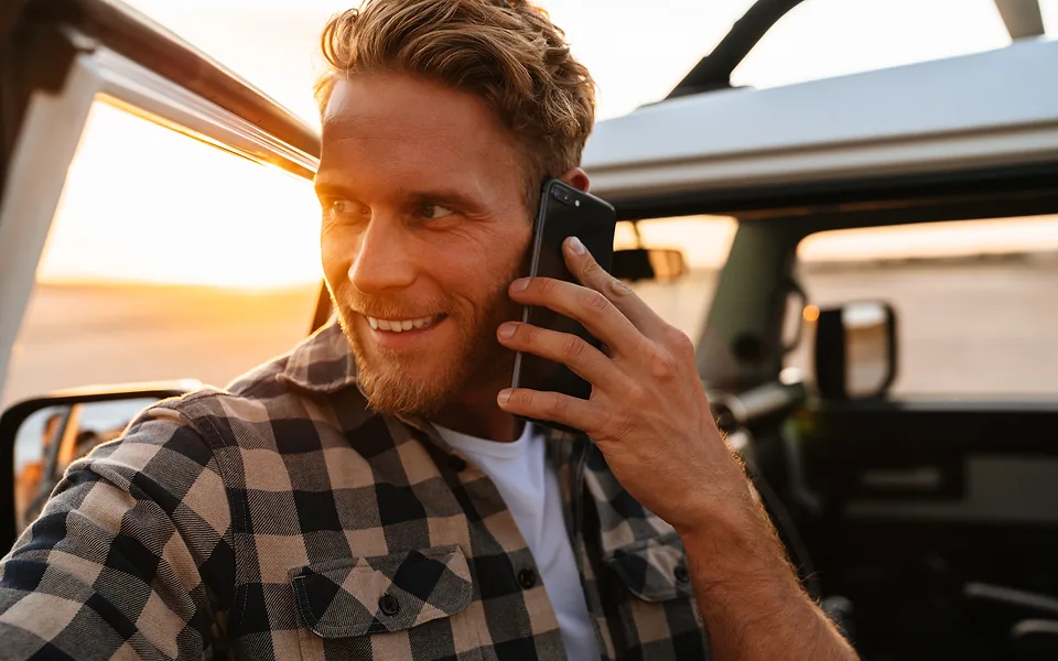 Smiling bearded man in a checked shirt taking a phone call beside an open-top vehicle at sunset