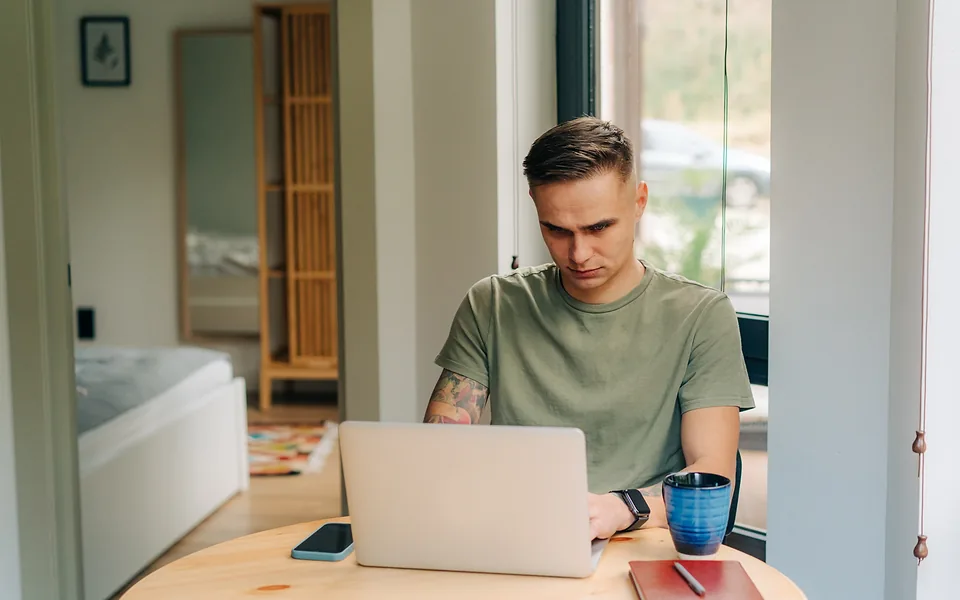 Man in a green t-shirt working on a laptop at a round timber table beside a window at home