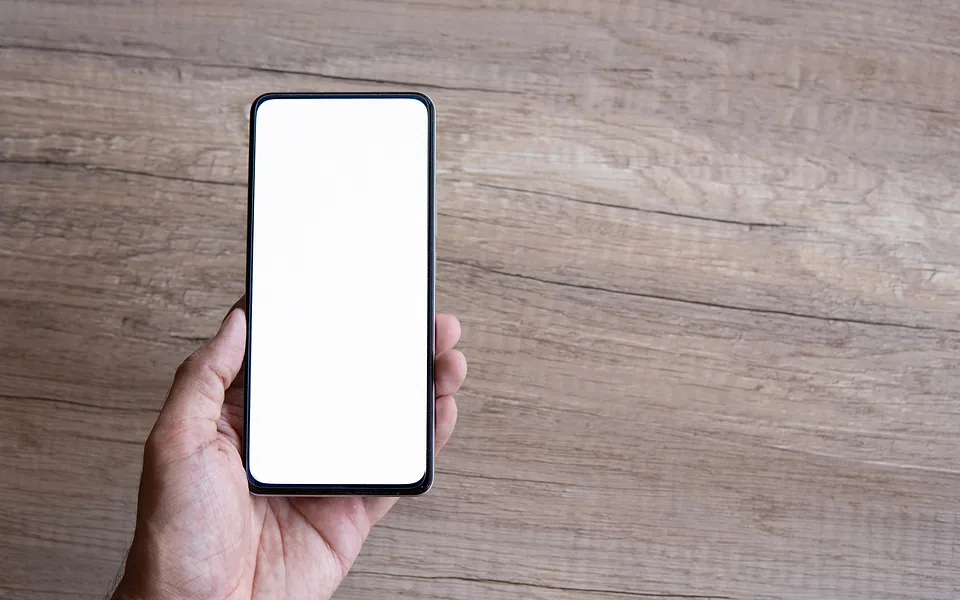 Hand holding a smartphone with a blank white screen above a grey timber tabletop