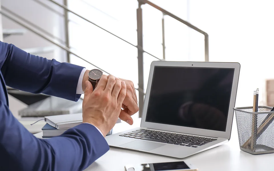 Man in a navy suit checking his wristwatch at a white desk with an open laptop in front of him